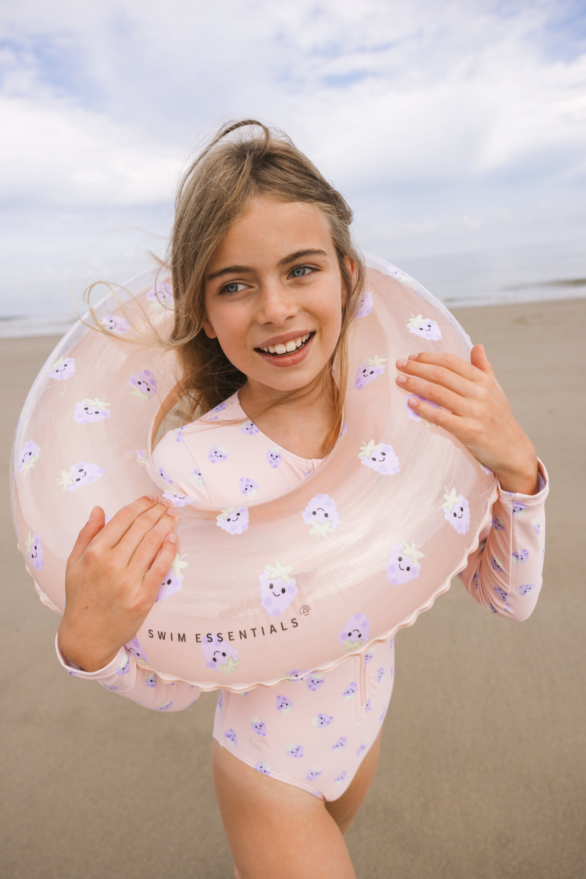 Smiling girl on the beach wearing a pink long-sleeve swimsuit with purple fruit print, holding a matching Swim Essentials inflatable ring around her shoulders, with the sea and sky in the background.