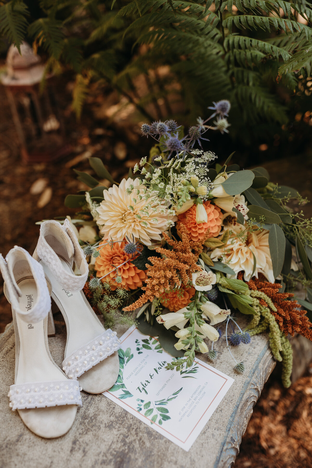 A colorful wildflower bouquet next to bridal shoes and a wedding invitation on a rustic wooden surface