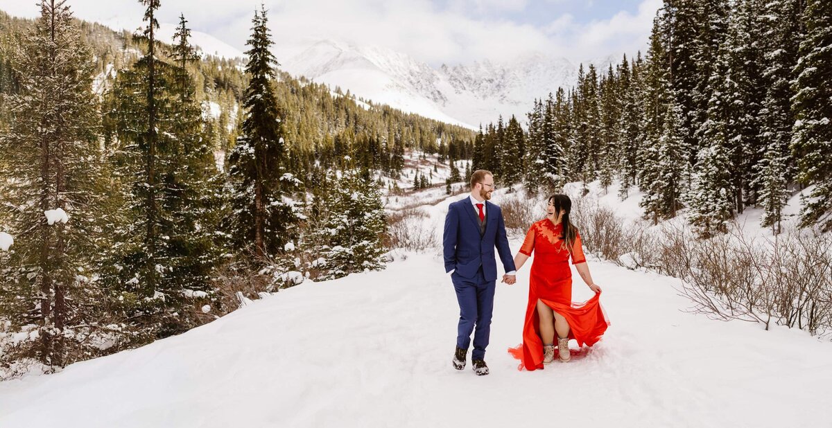 Bride in a red dress and groom in a blue suit holding hands and walking through fresh snow during their January winter elopement in Breckenridge, Colorado, surrounded by pine trees and mountain peaks