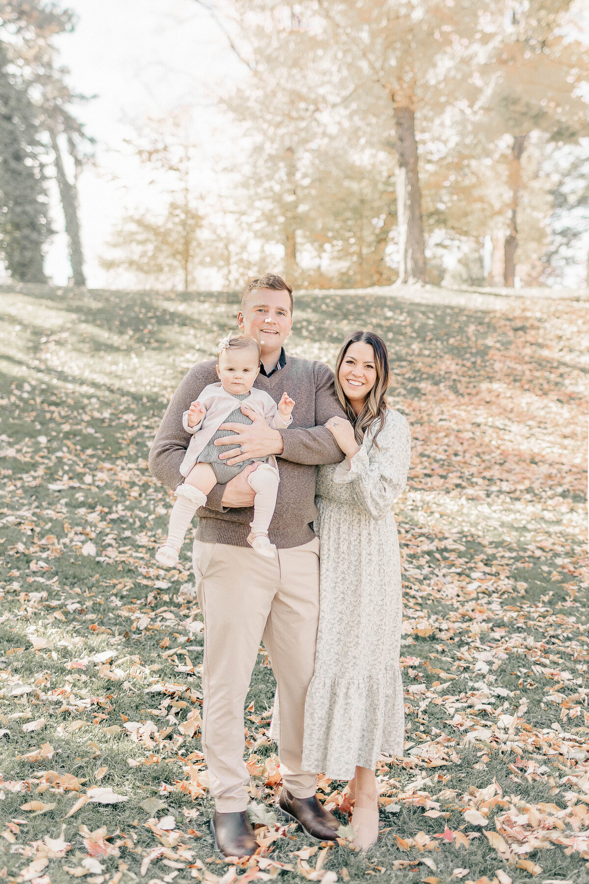 Family of three standing together on fallen autumn leaves at a Kansas City park during their baby girl’s first birthday photo session.