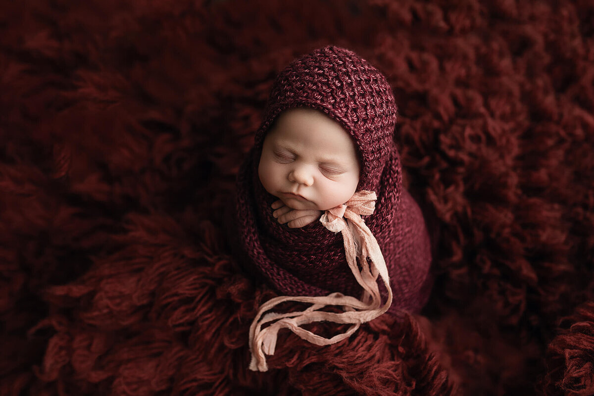 Newborn baby girl wrapped in a hand knitted maroon blanket wearing a matching bonnet.