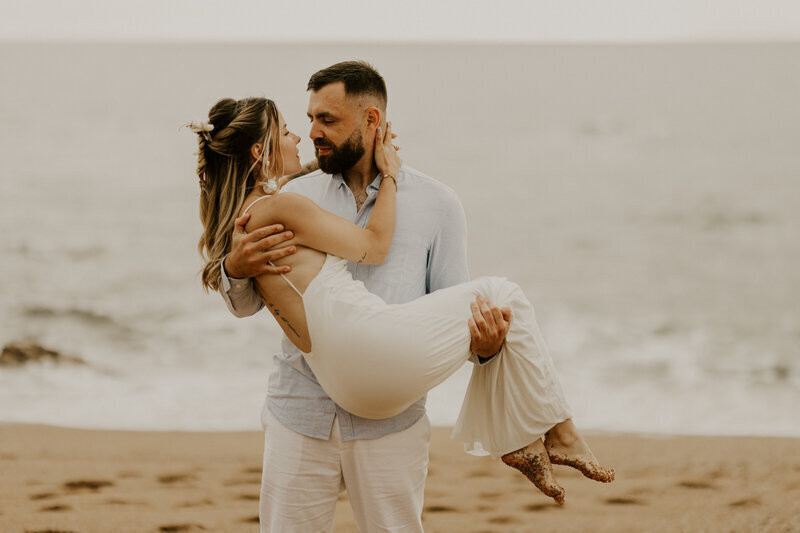 Homme en chemise bleu portant sa chéri dans ses bras lors d'un shooting photo de couple à la plage.