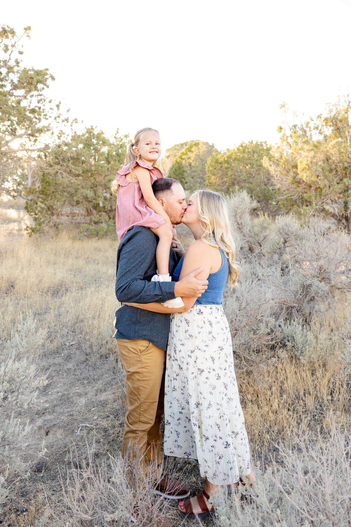 A little girl sits on her father’s shoulders, smiling brightly while her parents share a loving kiss below. The moment is filled with love and joy, captured in a beautiful natural setting.