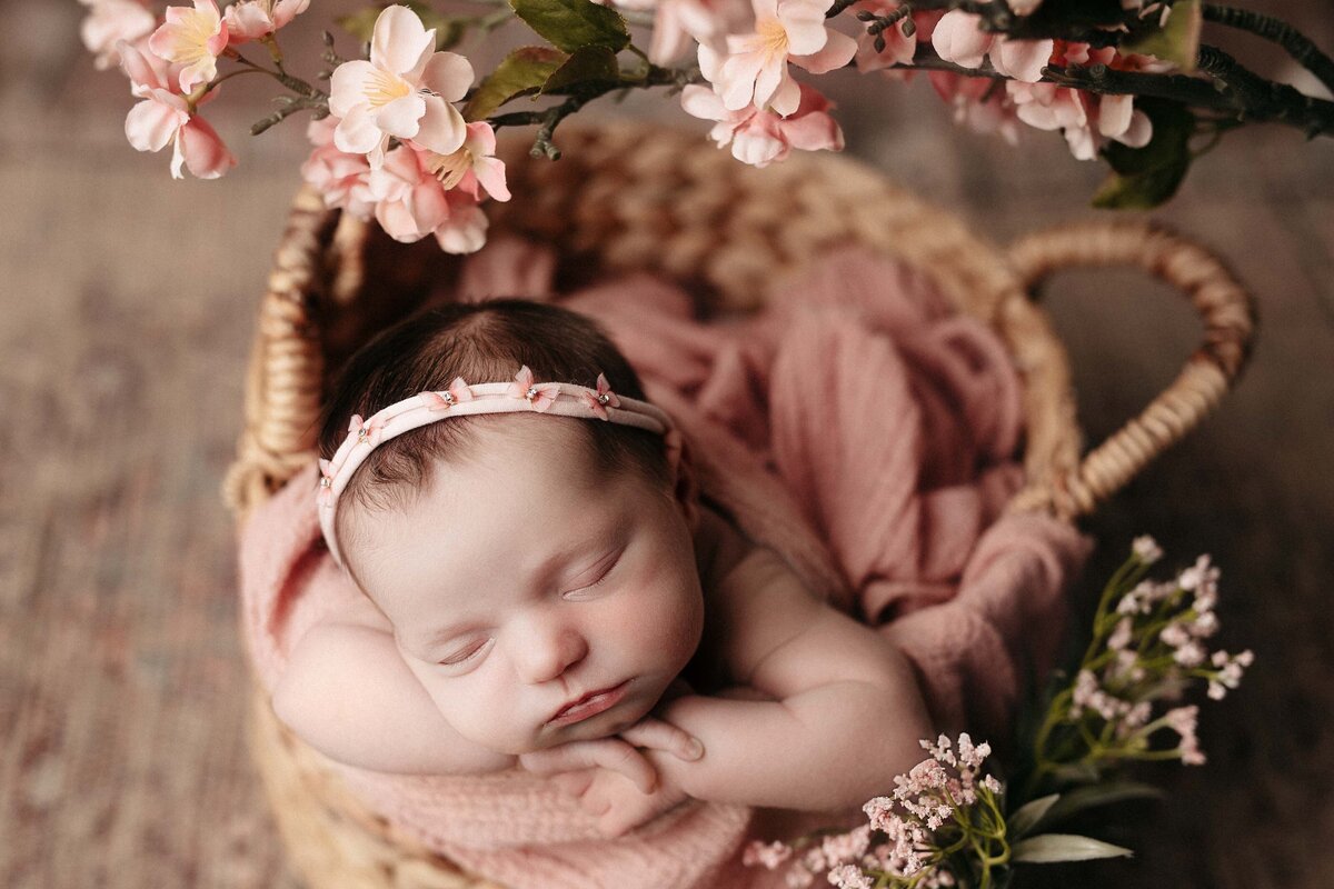 Studio Newborn Photography - Baby girl sleeping in woven basket with a pinky-apricot blanket. Surrounded by flowers in the foreground.