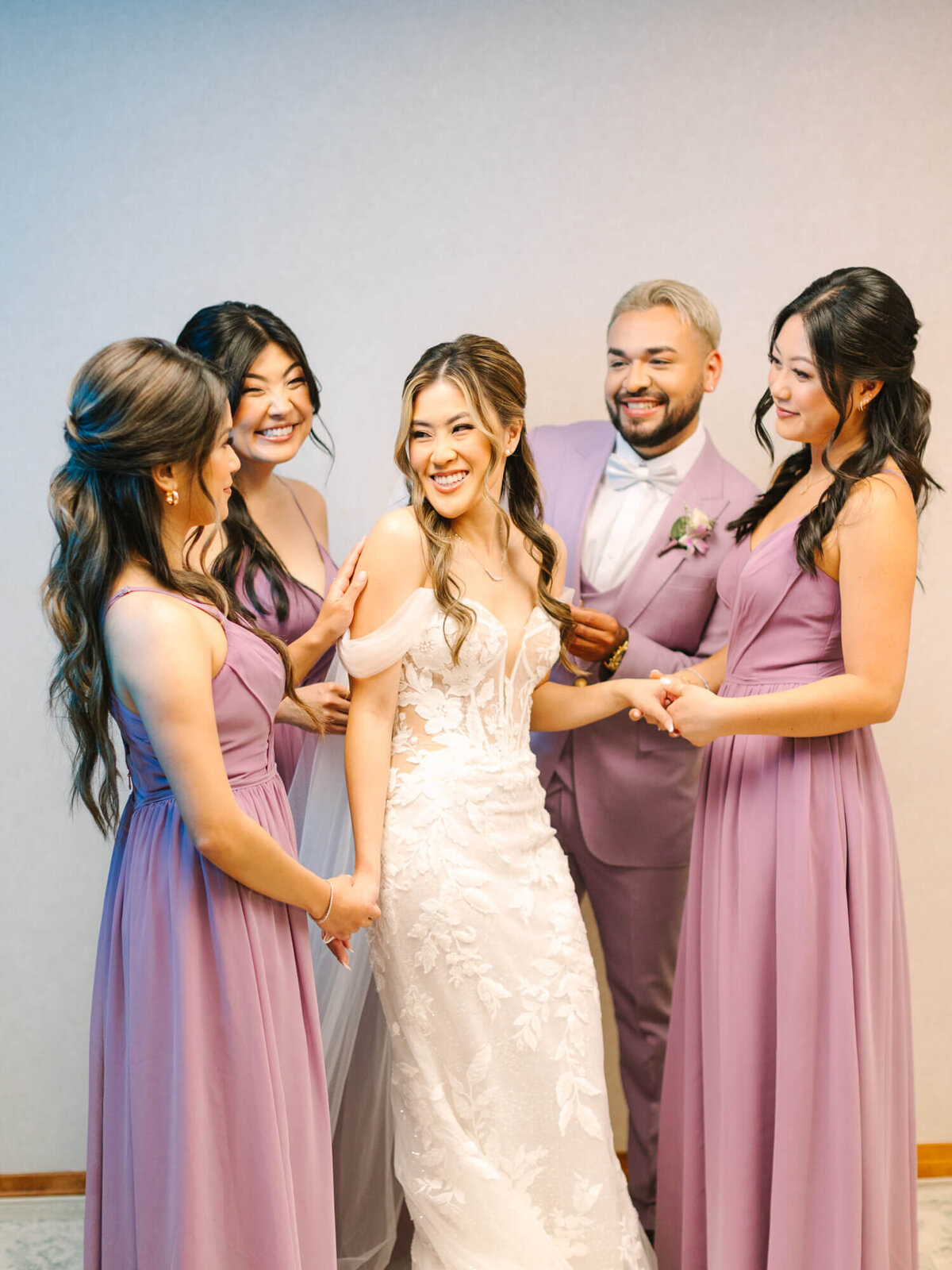 A joyful bride in a white lace gown is surrounded by three bridesmaids in lavender dresses and a groomsman in a mauve suit. They share smiles, conveying a sense of celebration and happiness.