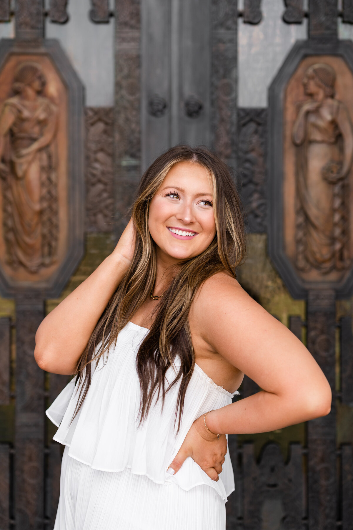 Texas A&M Senior girl smiling over shoulder with hand on hip and in hair while wearing white dress in front of the Administration Building front doors