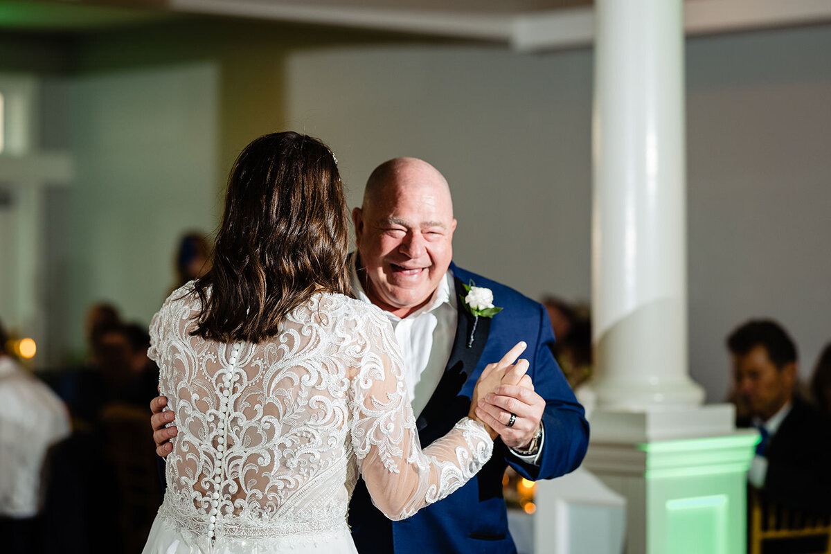 A bride in a lace gown dancing with a smiling man in a blue suit, enjoying a father-daughter dance.