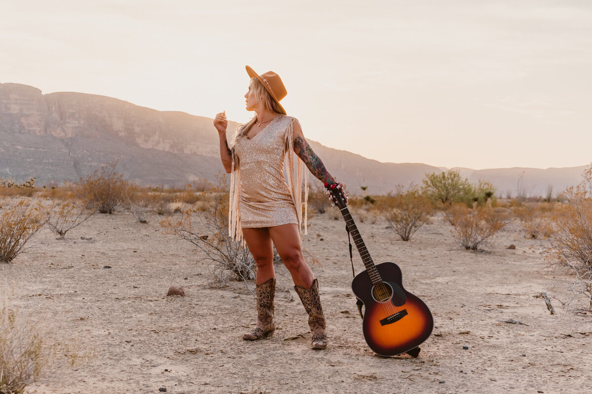 Woman standing in a desert landscape at Big Bend State Park, wearing a stylish hat and holding a guitar, embodying a free-spirited and adventurous vibe. Captured during a creative elopement session by ShutterUp Photography.