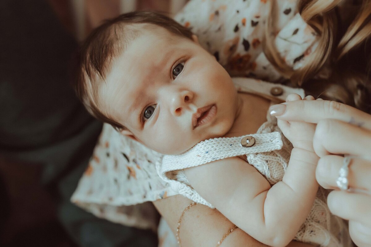 Baby held by an adult in a patterned shirt, looking up, with hand gently gripping a finger.