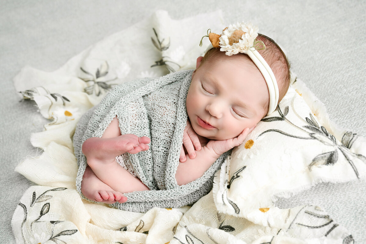 Newborn girl is sleeping on a daisy decorated blanket and pillow, smiling sweetly with one had under her cheek, crossing her little legs and wearing a headband with an adorable sunflower during her newborn session with the best newborn photographer in Pittsburgh, Katie Louise Photography.