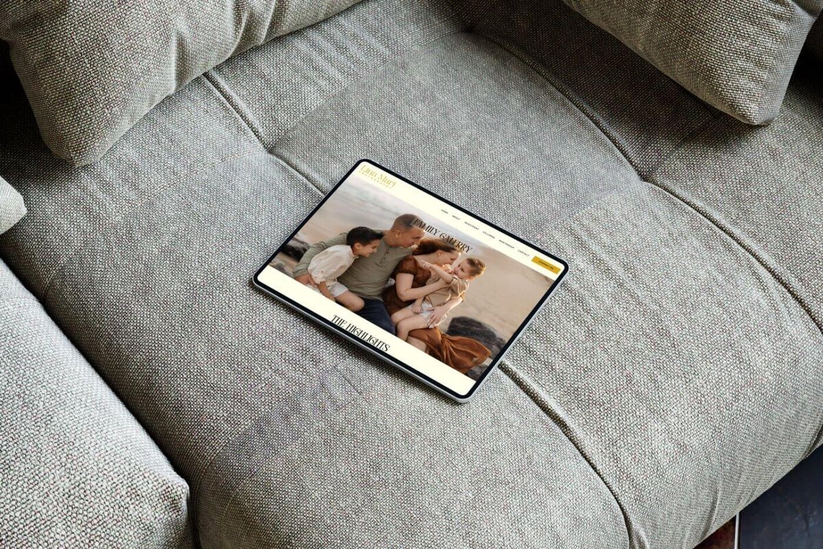 Tablet lying on a gray fabric sofa, open to a family photography website gallery featuring a couple cuddling their two young children during a beach photoshoot at sunset.