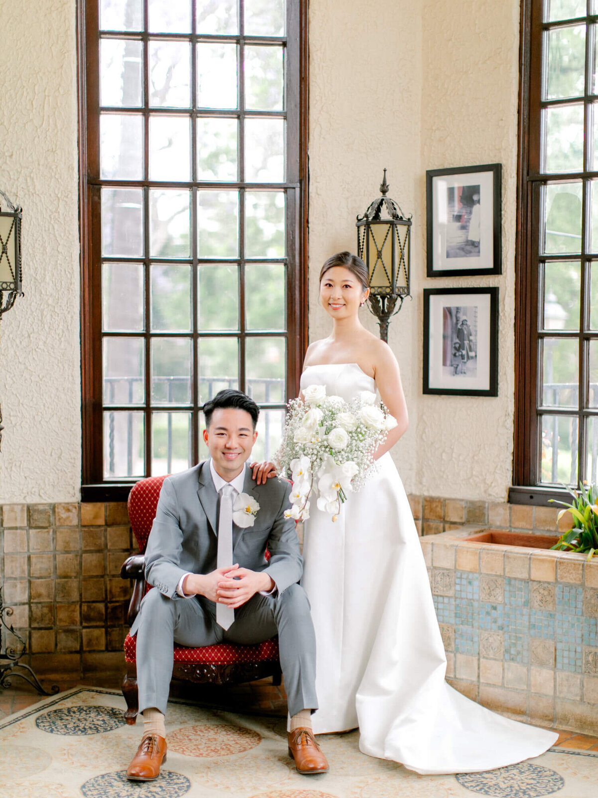 A bride in a white gown holds a bouquet, standing beside a groom seated in a grey suit. They are in a vintage room with mosaic tiles and large windows, exuding elegance and joy.