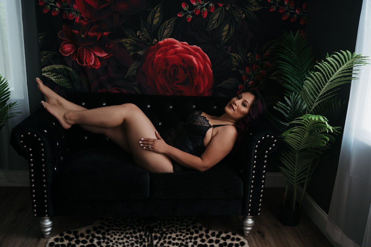 A woman in black lingerie lays across a black couch in front of a floral tapestry