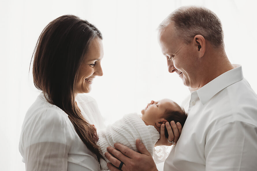 Mom and dad in white smile at their newborn baby in a white studio.