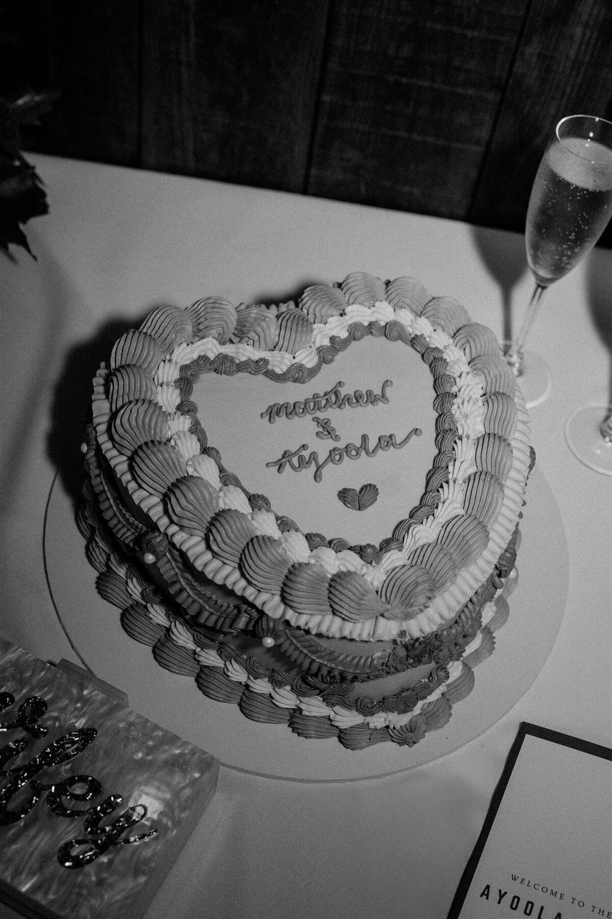 A vintage heart shaped wedding cake sits on a table. Champagne sits beside the cake.