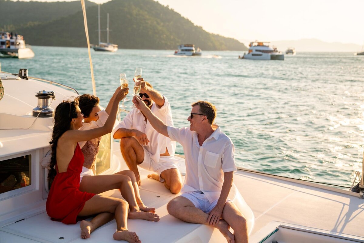 Four people are sitting on a yacht's deck, smiling and toasting with champagne glasses. The scene is set against a backdrop of blue water and distant mountains with other boats in view, under a bright, sunny sky.