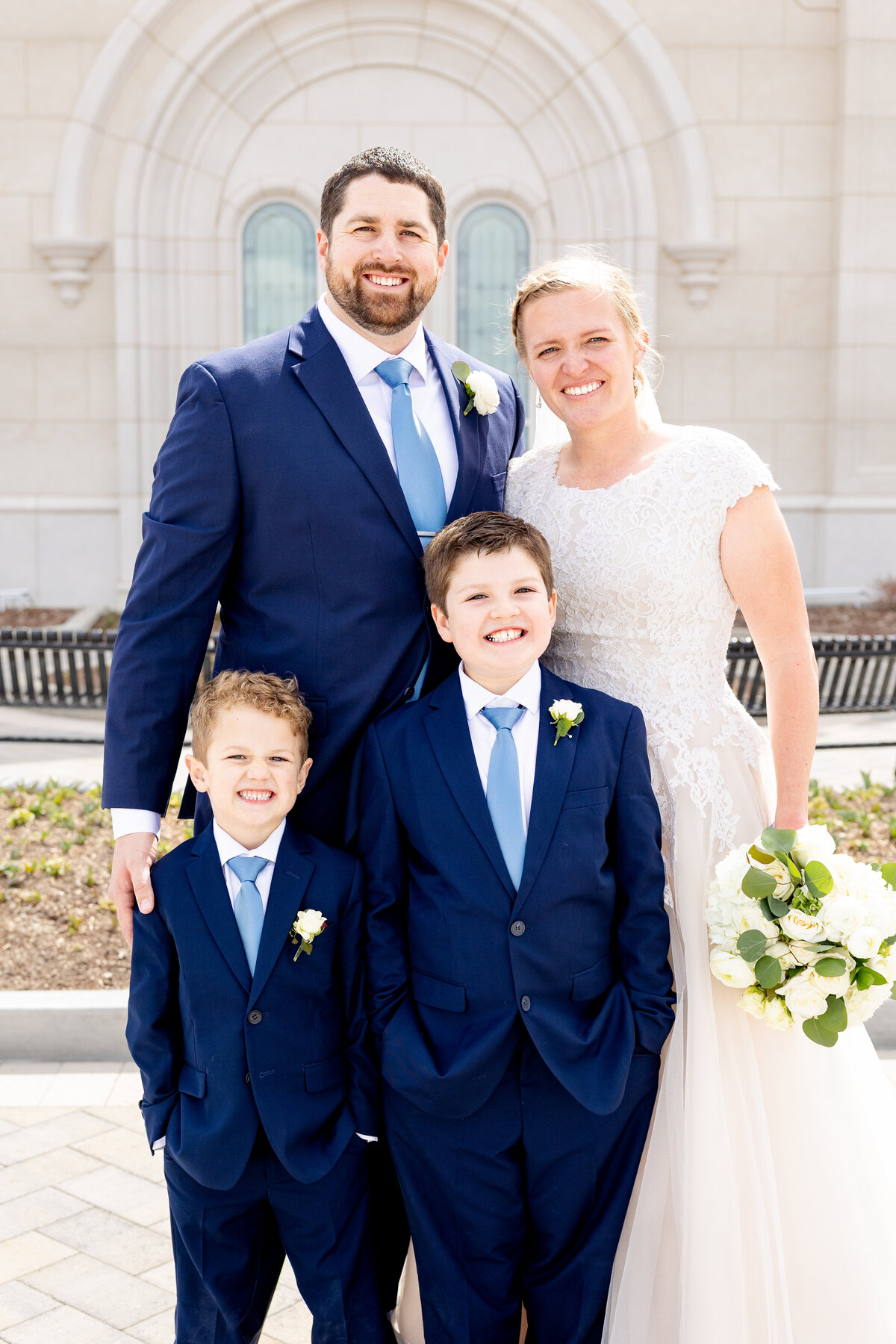 Bride and groom pose happily with their two sons dressed in matching navy suits and blue ties, standing in front of the temple.