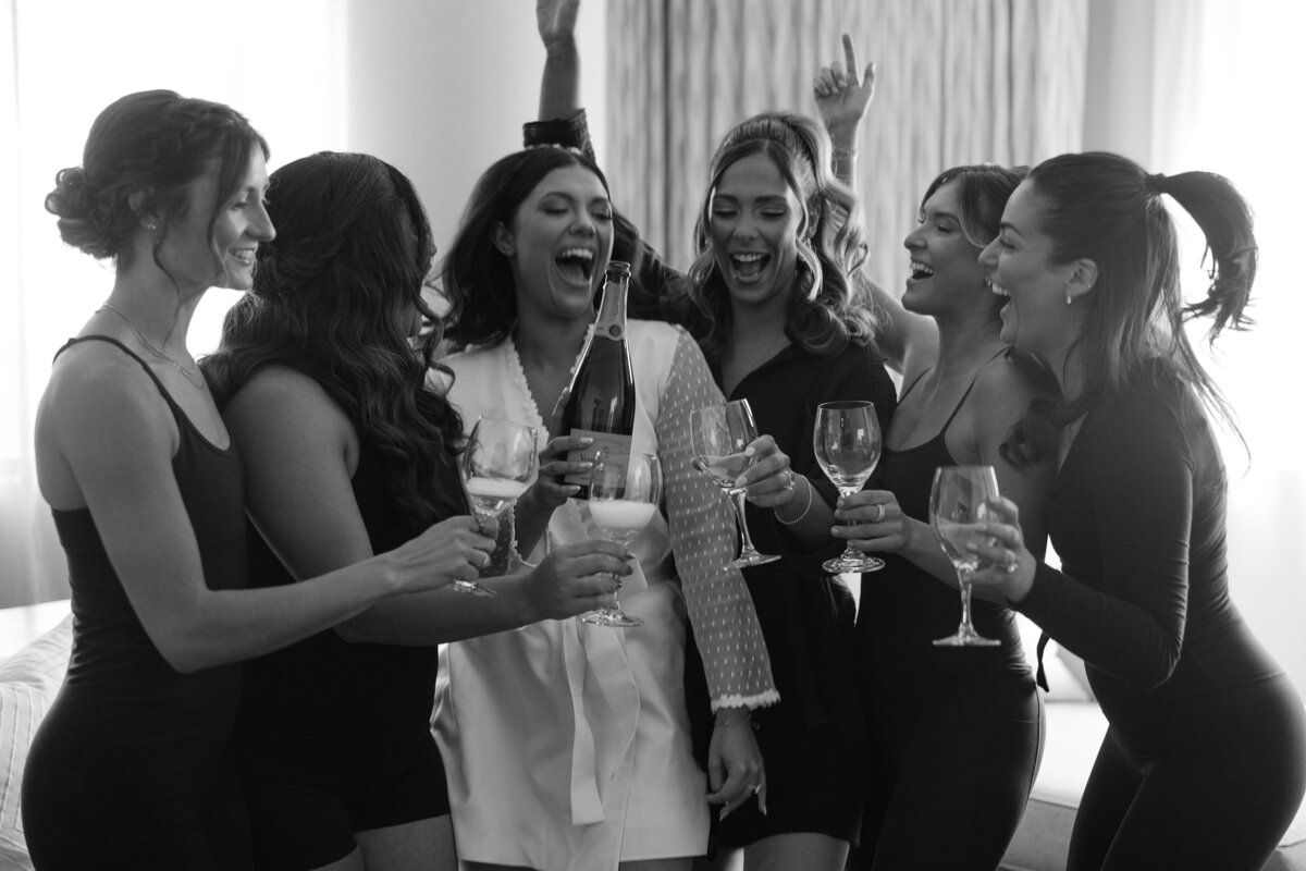 A black &  white image of a group of women mostly wearing black with the bride at the center wearing a white robe cheering as the bride holds a champagne bottle