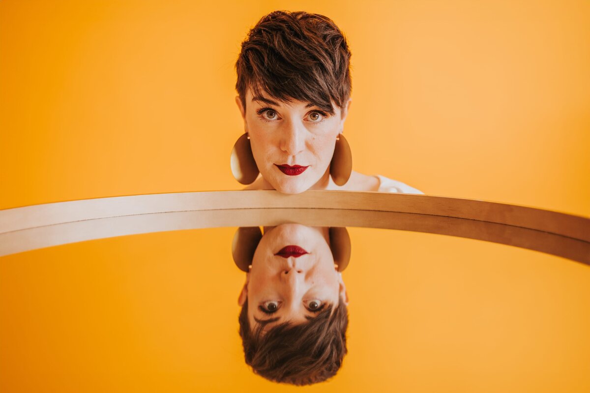Woman with very short brown hair wears large, gold earrings with her chin on a large mirror showing her reflection.