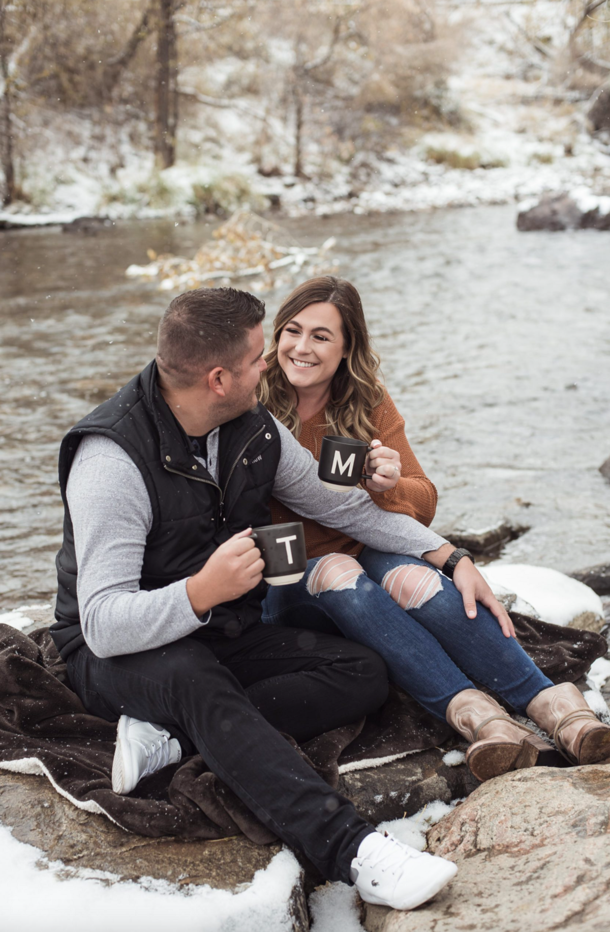 An engaged couple sits on the bank of a Colorado creek smiling at each other and sharing coffee in mugs with their initials.