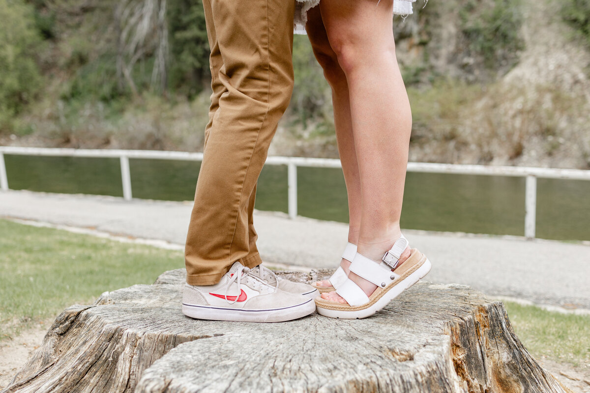 A couple stands on a large tree stump, showcasing their matching casual shoes, symbolizing their strong foundation as they begin their journey together in Vivian Park, Provo Canyon.