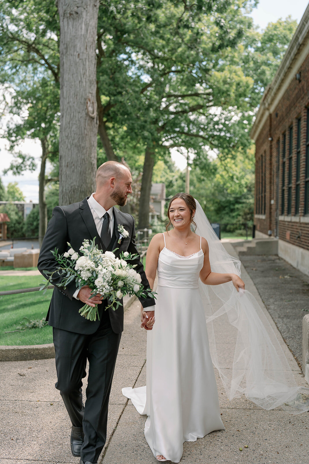 Bride and groom walking outside Leona Road while the groom carries the bridal bouquet during their elegant fall wedding in Grand Rapids MI.