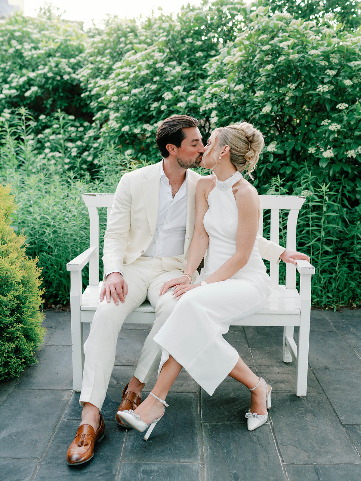 Couple wearing white as they kiss on a bench at salamander resort garden