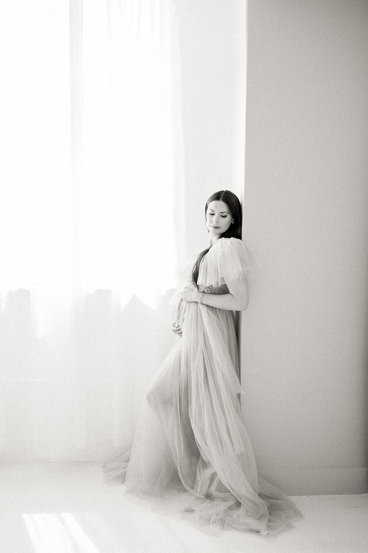 Black and white photo of an expecting Dallas mom leaning on a wall while she is dressed in a tulle maternity gown holding her belly in a Dallas photography studio.
