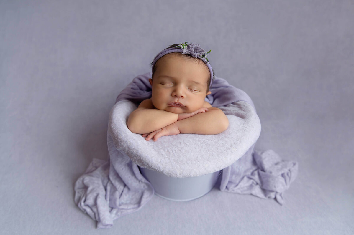 Newborn baby girl in a purple bucket on a purple background for her newborn photography session.
