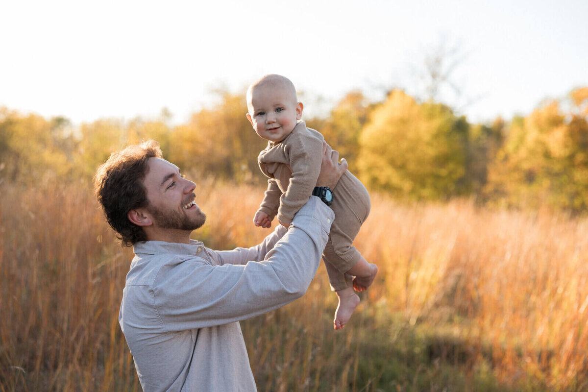 Father lifting his baby joyfully in an outdoor setting with autumn trees and tall grass in the background.