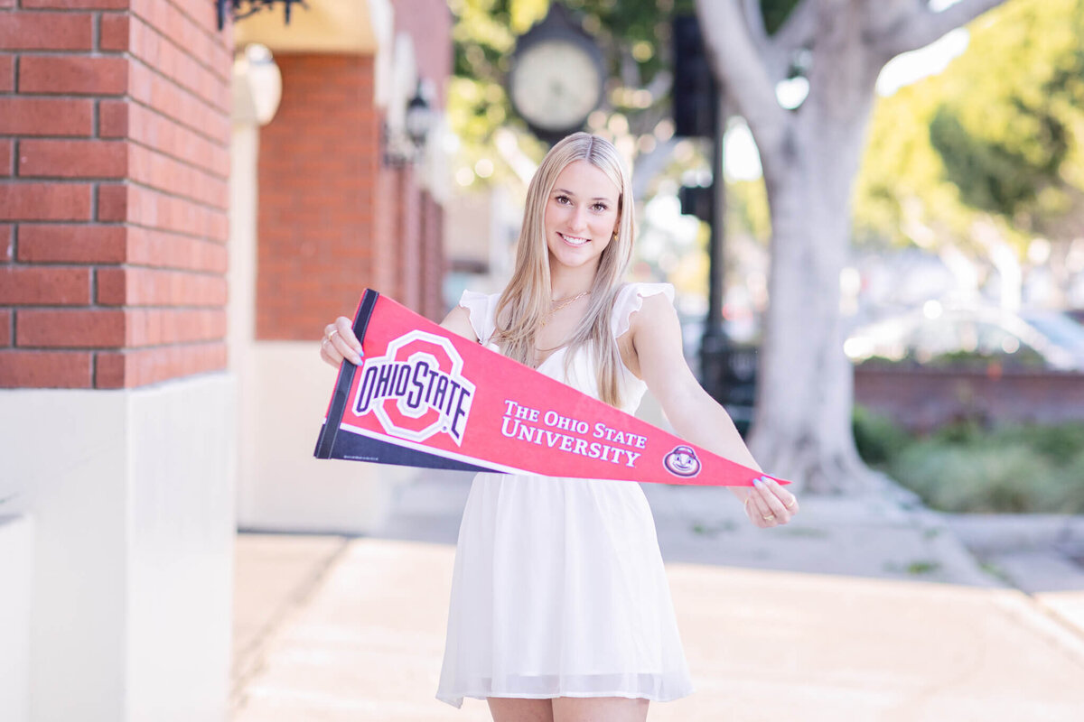 A person in a white dress stands outdoors, holding an Ohio State University pennant. The background features a brick wall and a large clock. Trees and a sunny sky are visible.