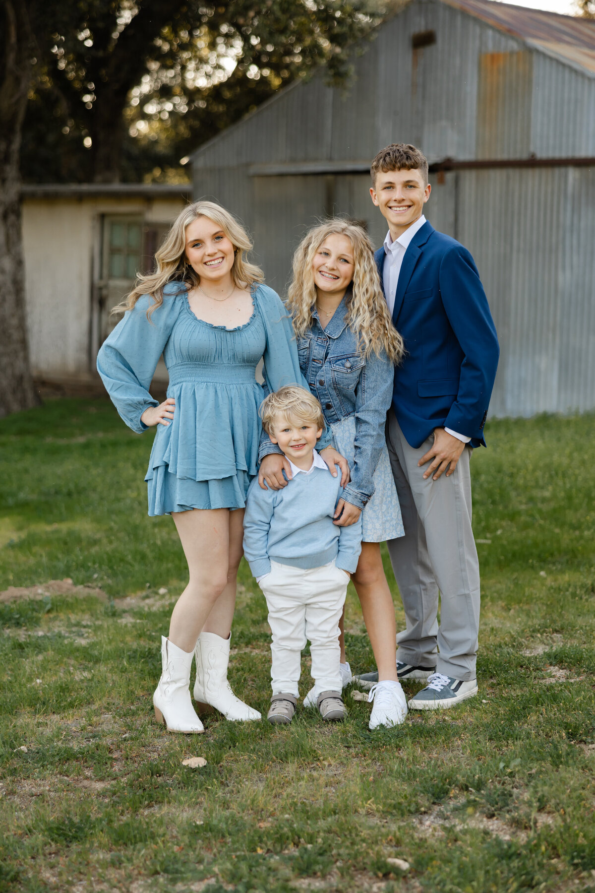 Four siblings standing together in front of rustic barn backdrop, smiling in coordinated outfits. Photographed by Yucaipa family photographer Kaitlyn Dawn Photography.