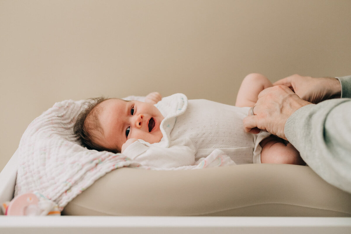 A father dresses his newborn daughter as she lays on her changing table, she is yawning and looking at the camera