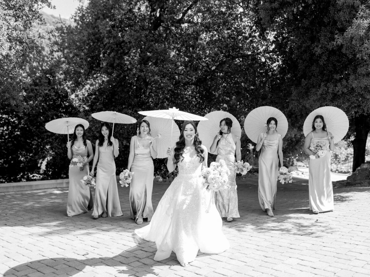 Bride in white gown smiles with six bridesmaids in gowns, holding parasols and bouquets, walking on a sunlit path, framed by lush trees. Elegant and joyful.