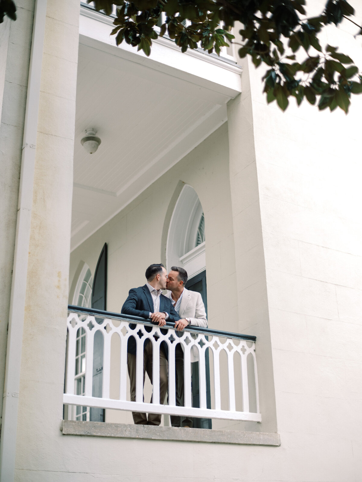A couple shares an affectionate kiss on a charming balcony, encapsulating a candid moment of romance and intimacy in a beautiful architectural setting during a serene day.
