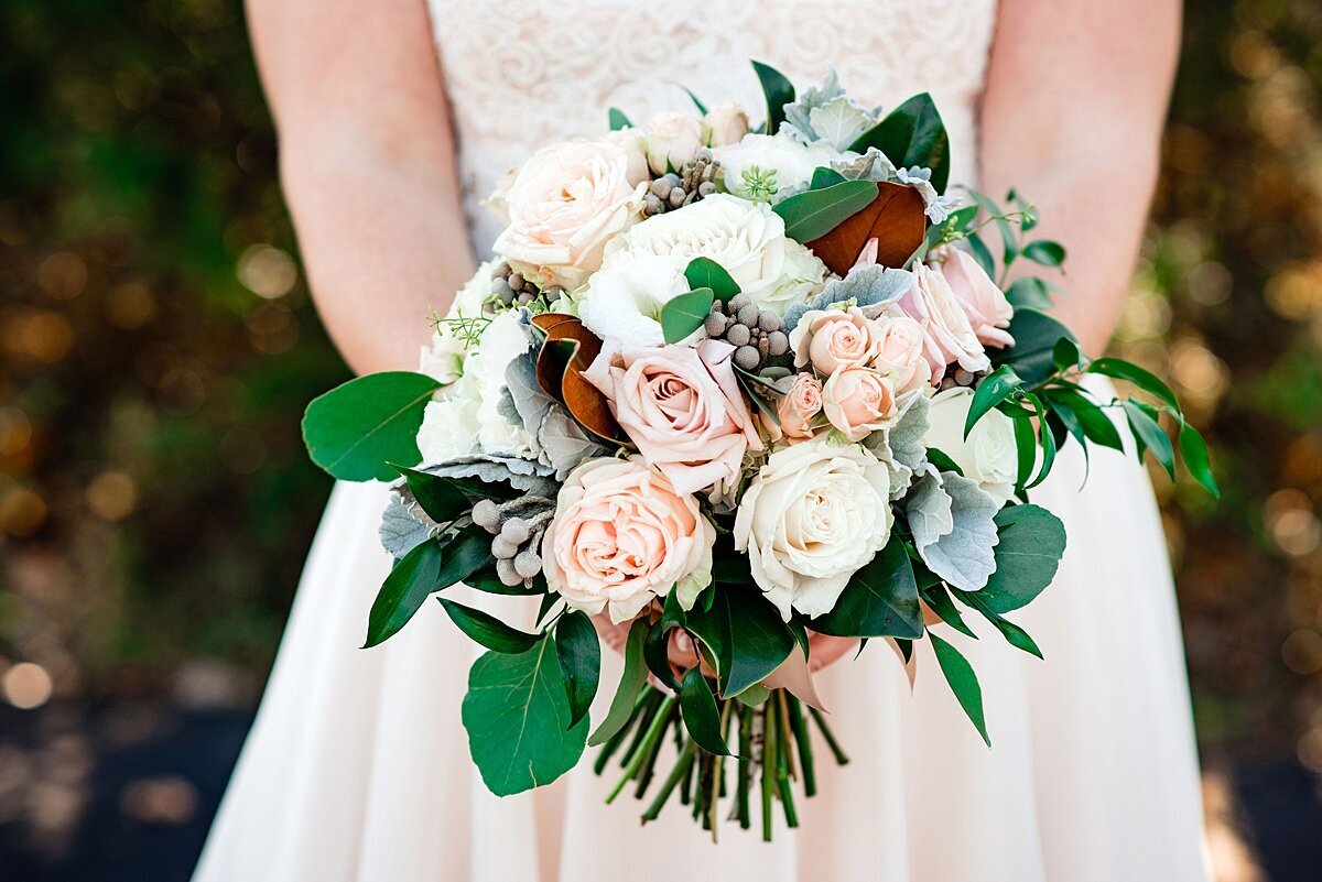 Large round bouquet of vlush, ivory and white roses with burgundy and soft gray accents, greenery and magnolia leaves for a fall wedding at Sycamore Farms