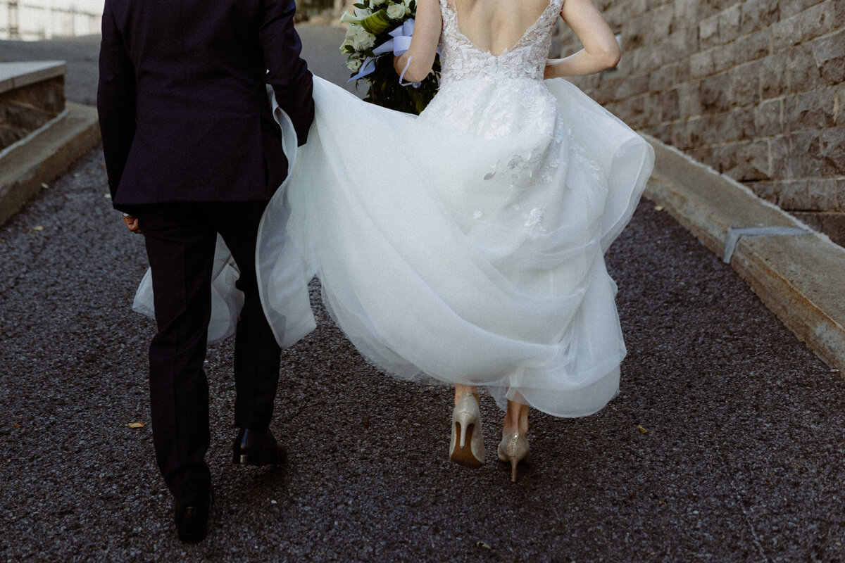 closeup-of-a-groom-holding-his-brides-dress-as-they-walk-up-a-hill-away-from-the-camera-1