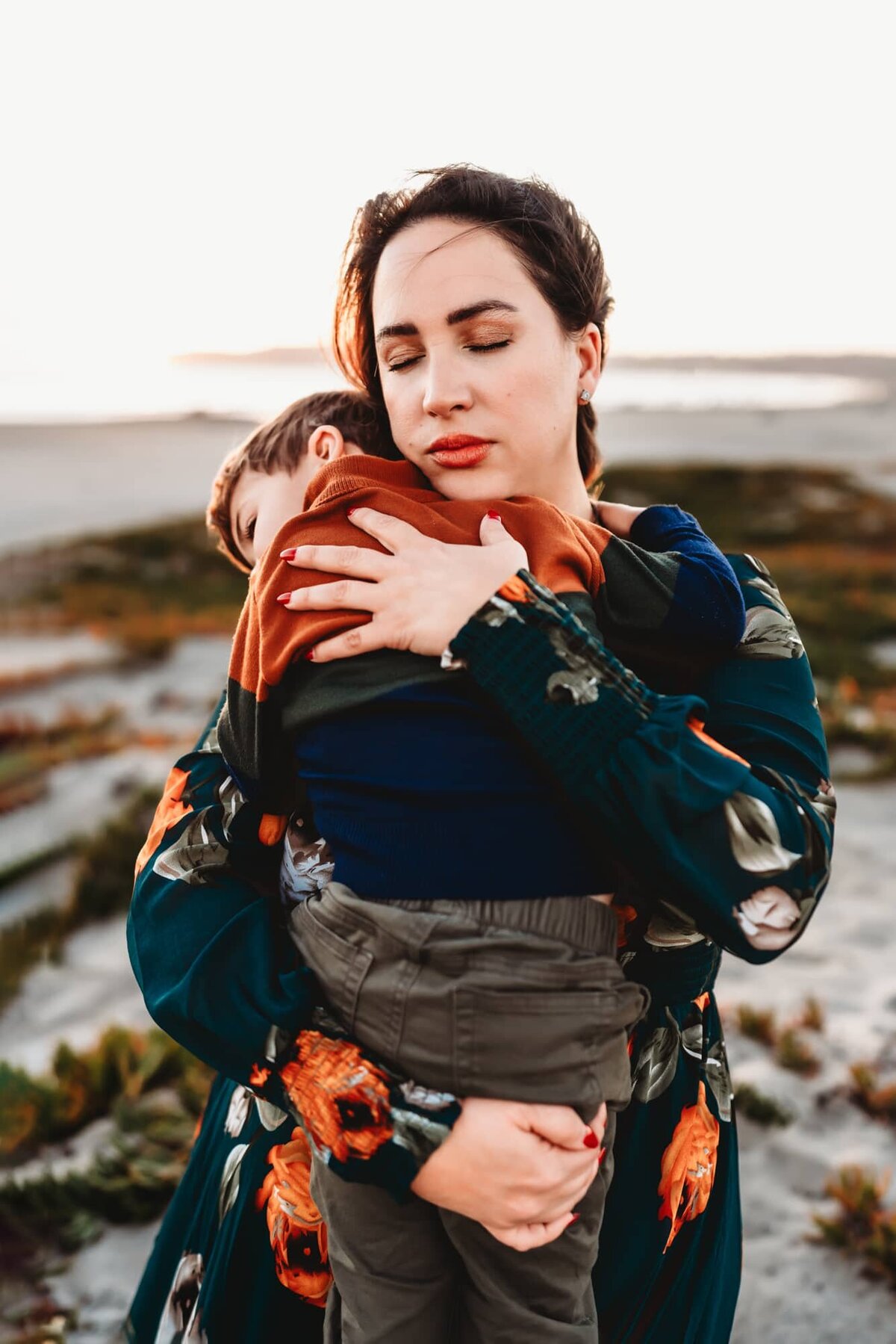 A mother  holds her toddler son in her arms on a plant covered sand dune on Coronado Island, during a beach family photo shoot by Love Michelle Photography
