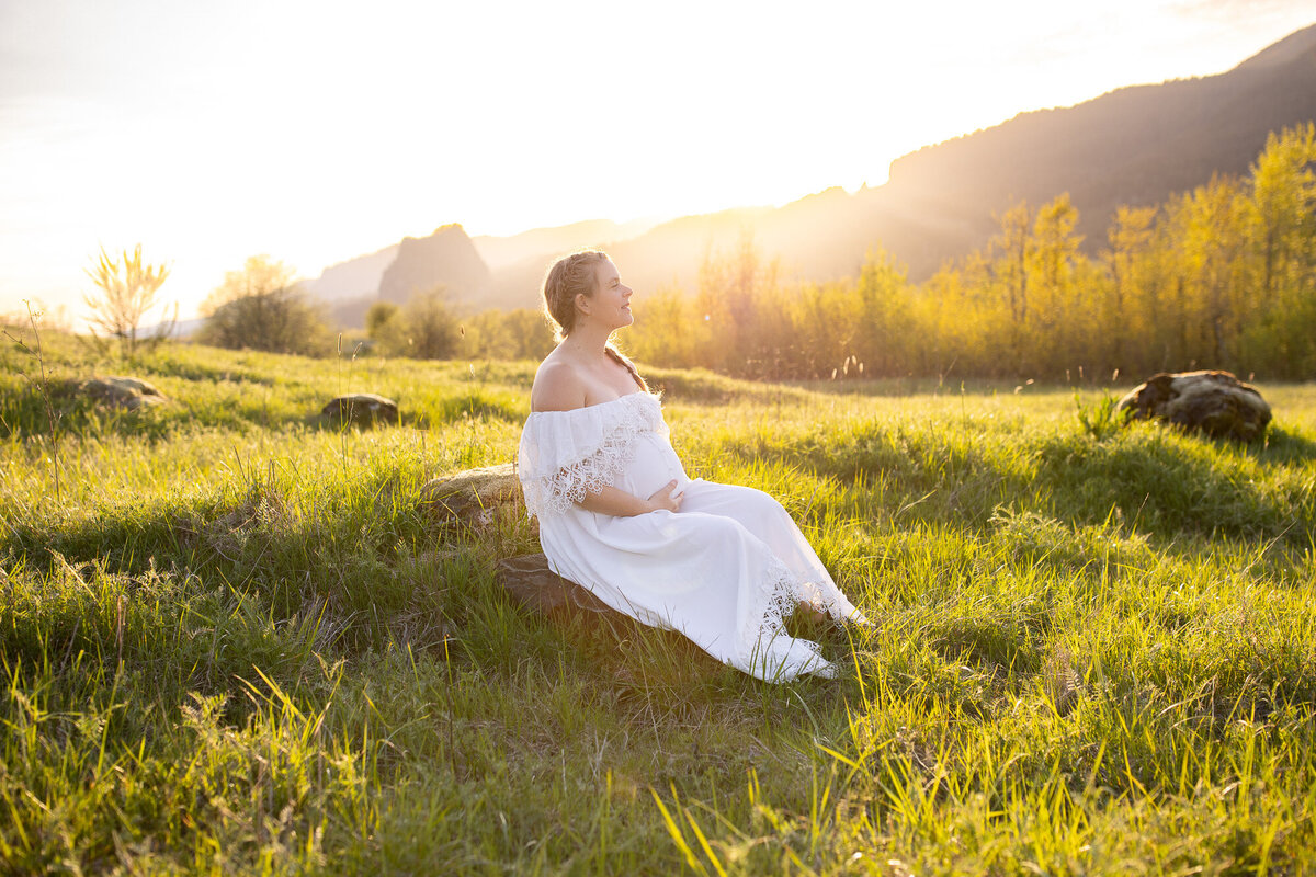A pregnant woman in a white long dress with lace looks up at the sky at sunset in front of Beacon Rock. 