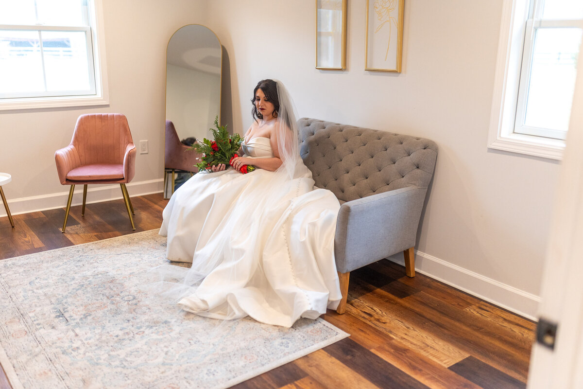 In a serene Leesburg VA wedding venue, a bride in a white gown sits gracefully on a gray couch, holding her bouquet. Beside her, the veil cascades gently. The room's elegance is marked by framed art on cream walls and a patterned rug covering the wooden floor.