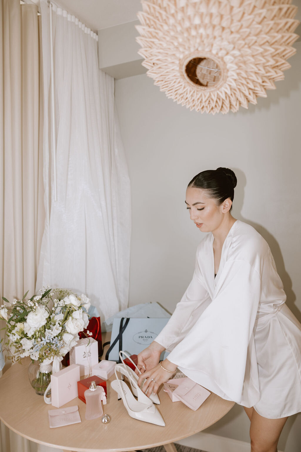 bride in silk white robe reaching towards her prada shoes on a table with flowers and other accessories in a hotel room