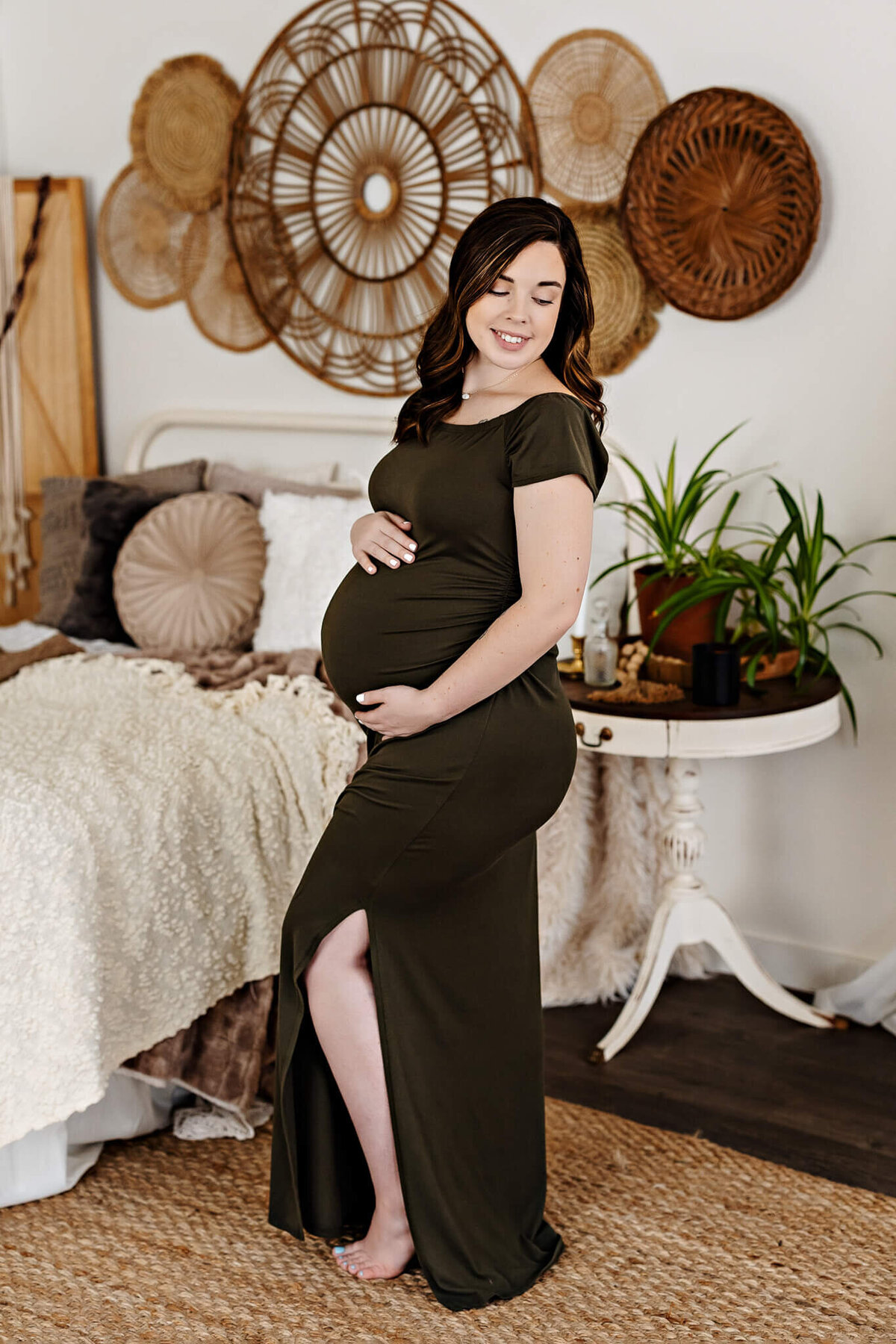 A smiling pregnant woman in a dark green dress stands barefoot beside a bed in a cozy room adorned with woven wall art and indoor plants. She gently cradles her belly, beautifully captured by a Bloomington maternity photographer.