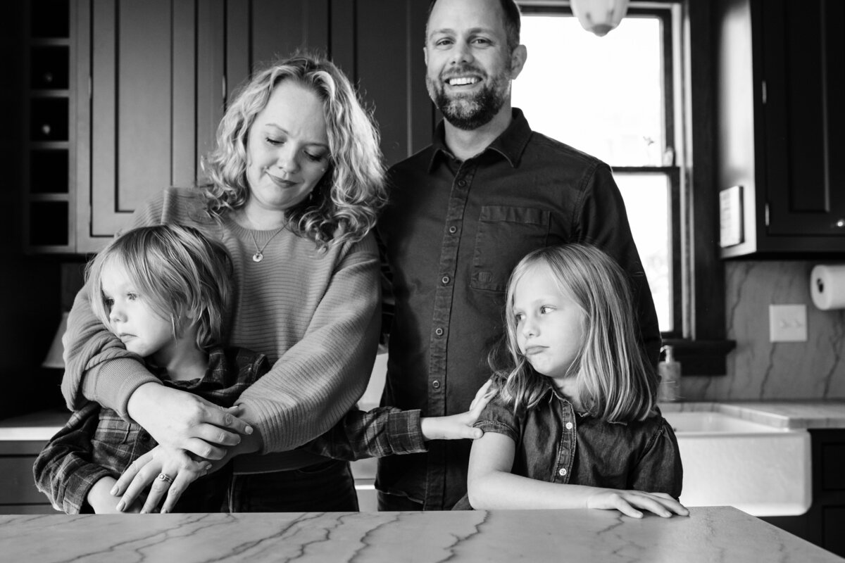 A black and white photo of a family standing together in a kitchen. The mother is holding one child close, who looks away, while the father stands smiling with the other child gently touching the mother's arm.