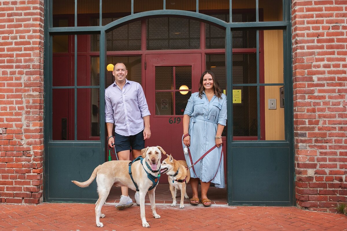 happy-couple-with-their-labrador-and-shiba-inu-mix-posing-in-front-of-a-red-bricked-historic-building-in-the-holy-cross-neighborhood-of-indianapolis-during-a-pet-friendly-photoshoot