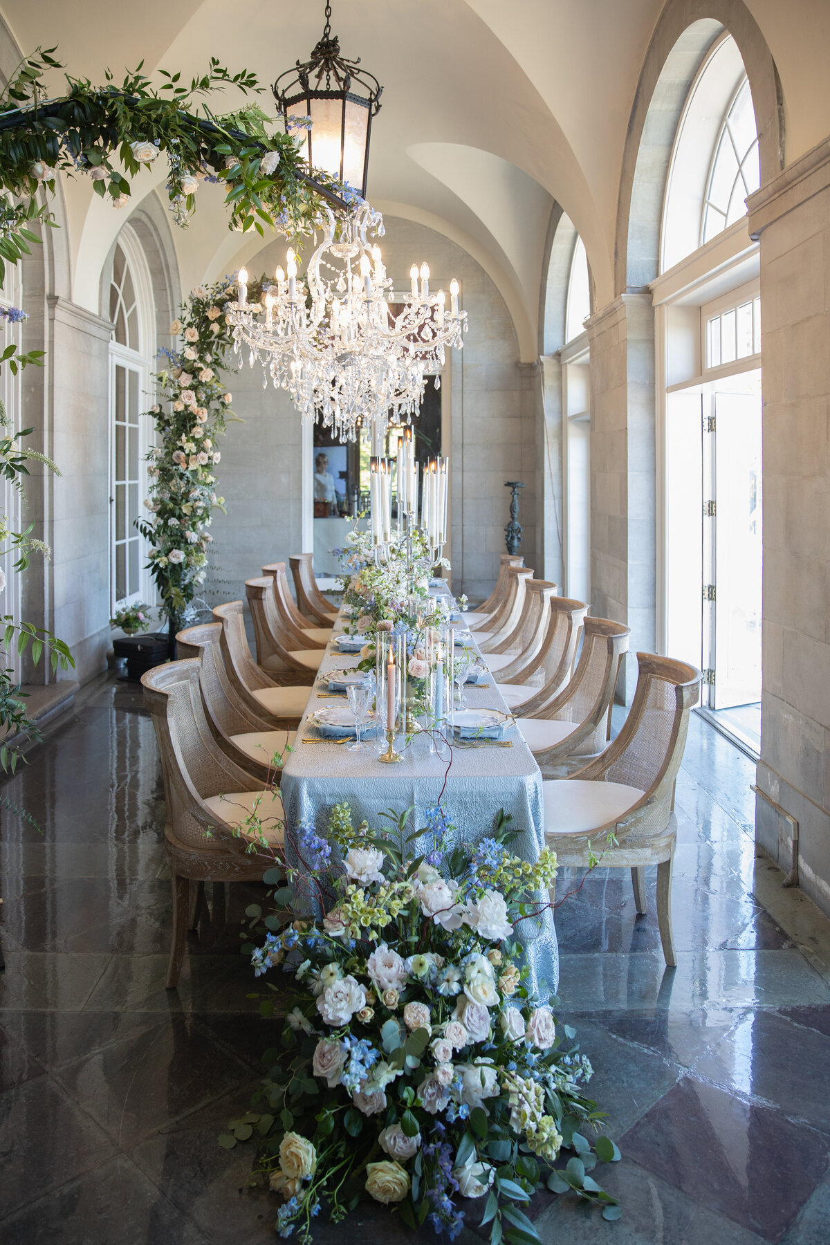 Aldrich Mansion Wedding Photos | Kelsey Sheehan Photography Timeless Rhode Island Weddings | Elegant dining room with a long table set for a banquet, adorned with blue and white flowers and a silver tablecloth. A crystal chandelier hangs above, creating a luxurious atmosphere.