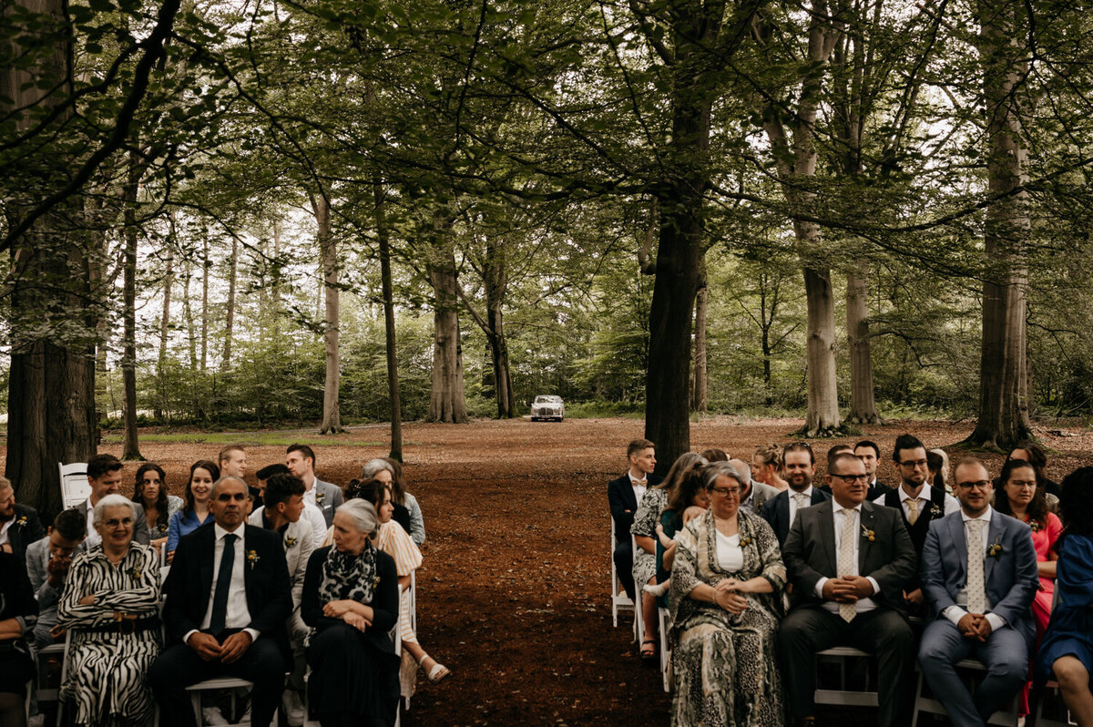 De trouwauto arriveert bij de ceremonie in het bos, met een romantische setting tussen de bomen.