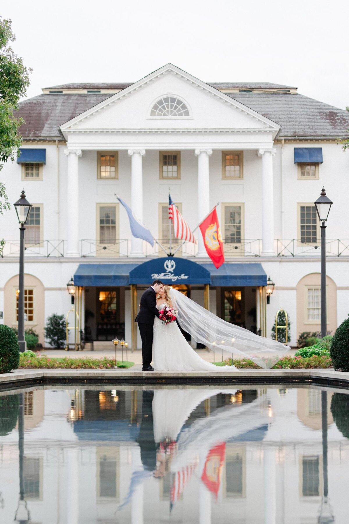 A couple stands by a reflective pool in front of an elegant building, with the bride's long veil flowing behind them.