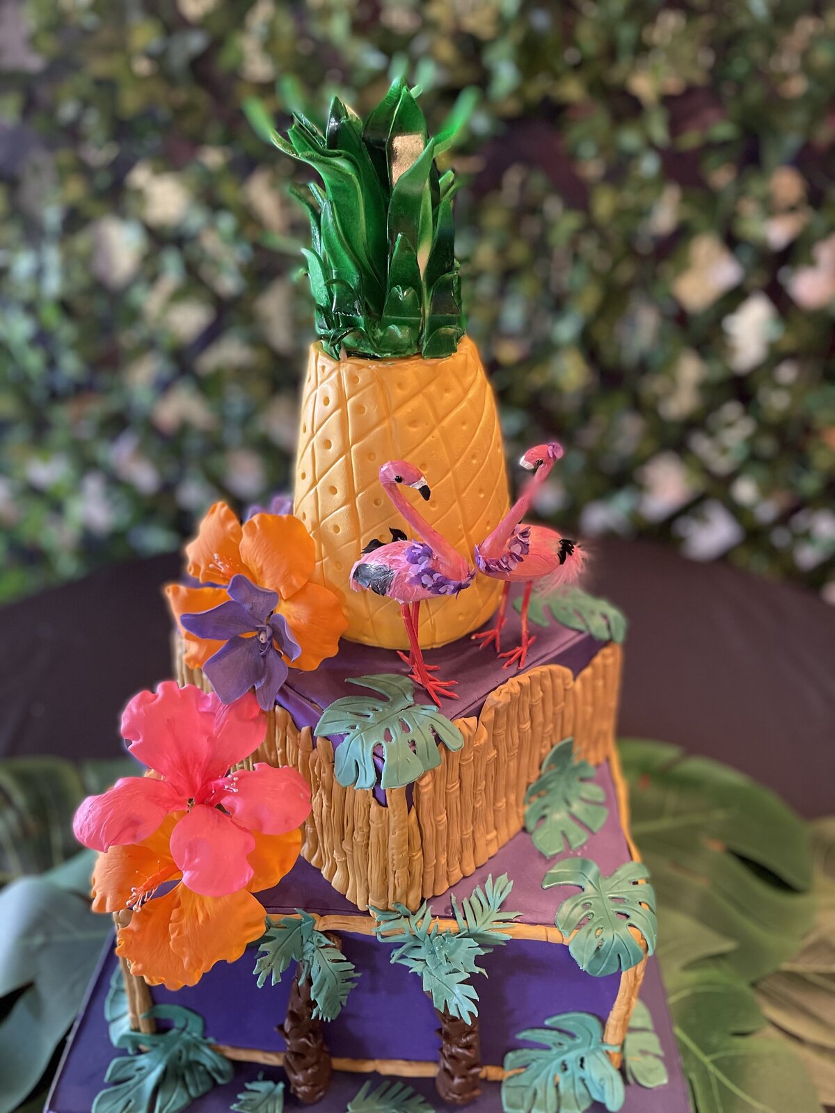 Tropical theme 3 tier wedding cake. Bottom tier is deep purple square with Monestera leaves, plam treese and bamboo edges; middle tier is square with bamboo  and cascading pink and orange hibiscus ; purple call lilies, purple orchids dnd pink flamingos. Top tier is carved pineapple.