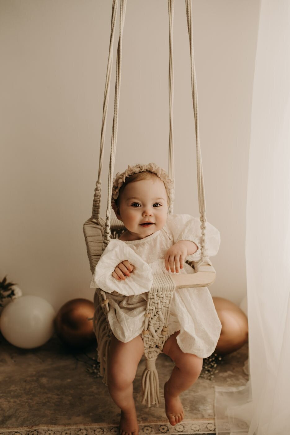 A cute baby sitting on a swing and smiling and very happy in a white dress with balloon in the background