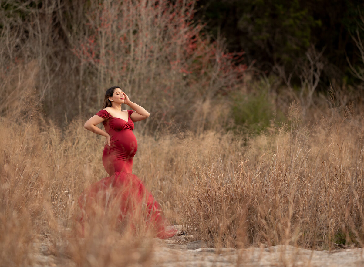 Glamorous outdoor maternity portrait of woman in red gown surrounded by golden tall grass near Leander, Texas